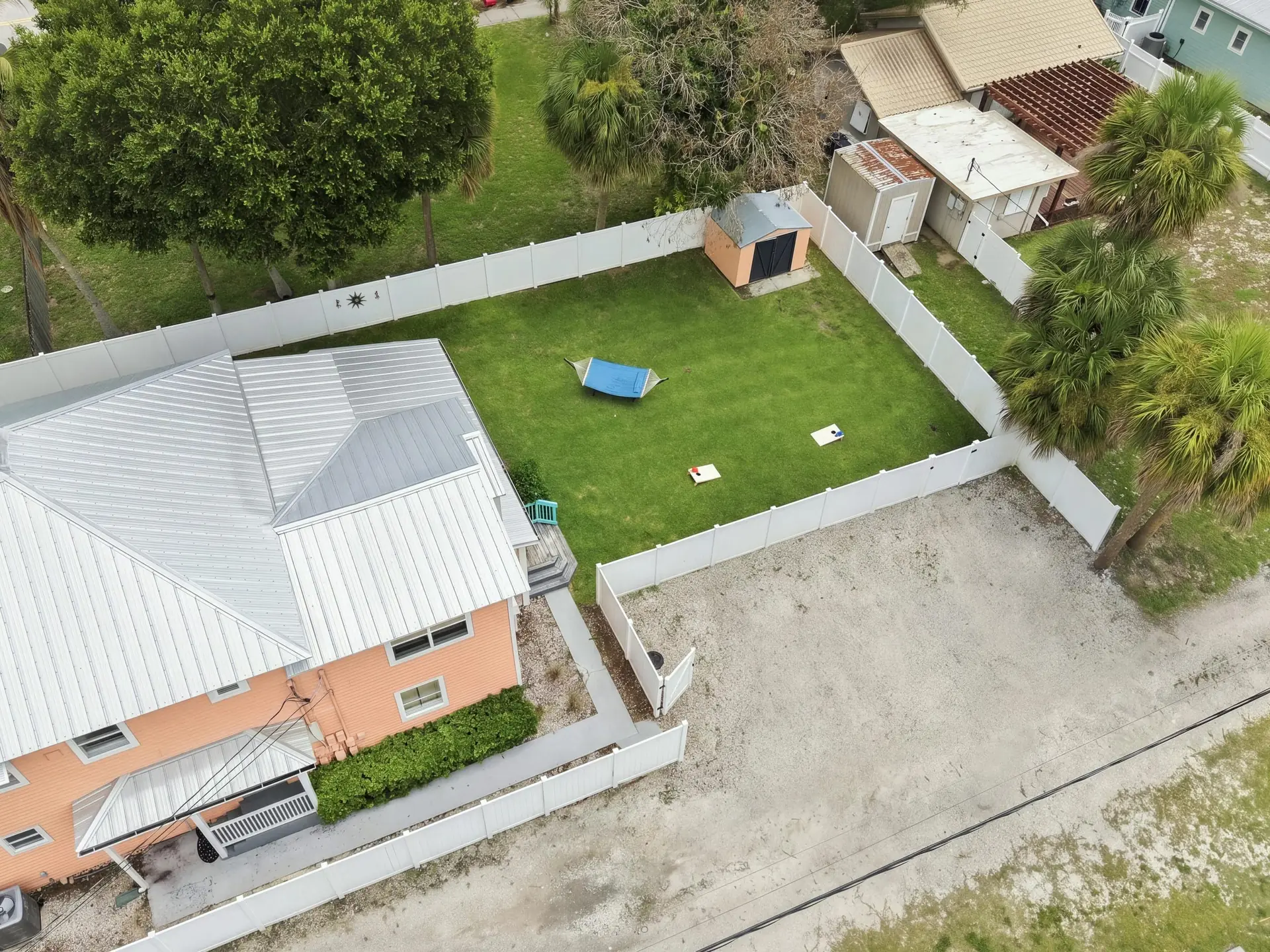 Overhead aerial view of the fenced backyard showing the blue hammock, corn-hole boards, storage shed and the home’s metal roof