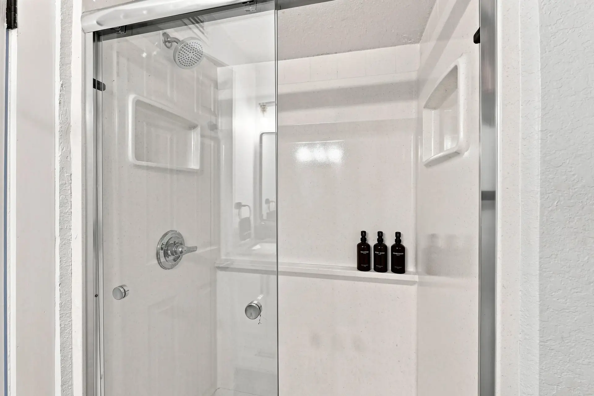 Walk-in shower with a glass sliding door and refillable dark amber toiletry bottles on the built-in ledge