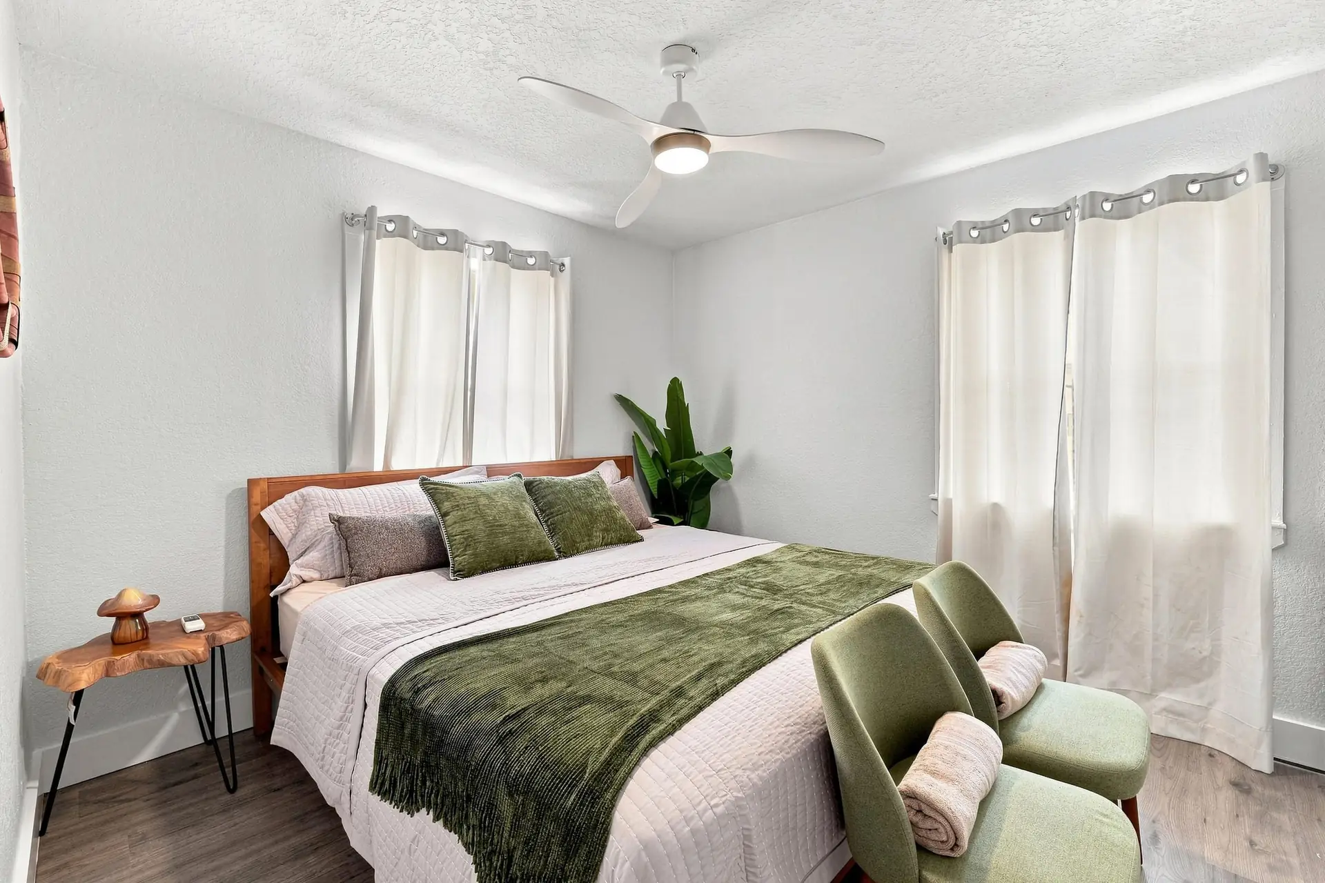 King bedroom with a warm wood headboard, sage green velvet throw and matching sage armchairs between two curtained windows
