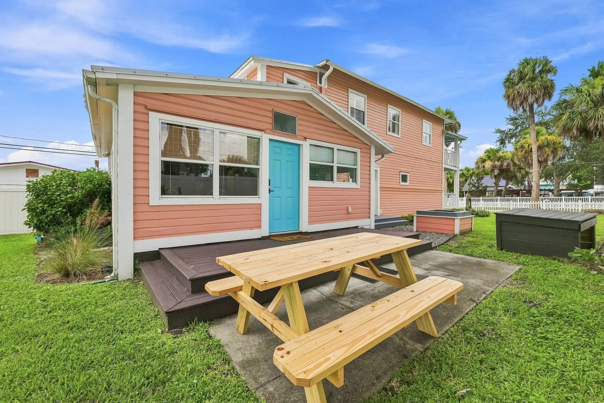 Back of the home with a turquoise door, a new wooden picnic table on the patio, raised garden beds and palm trees