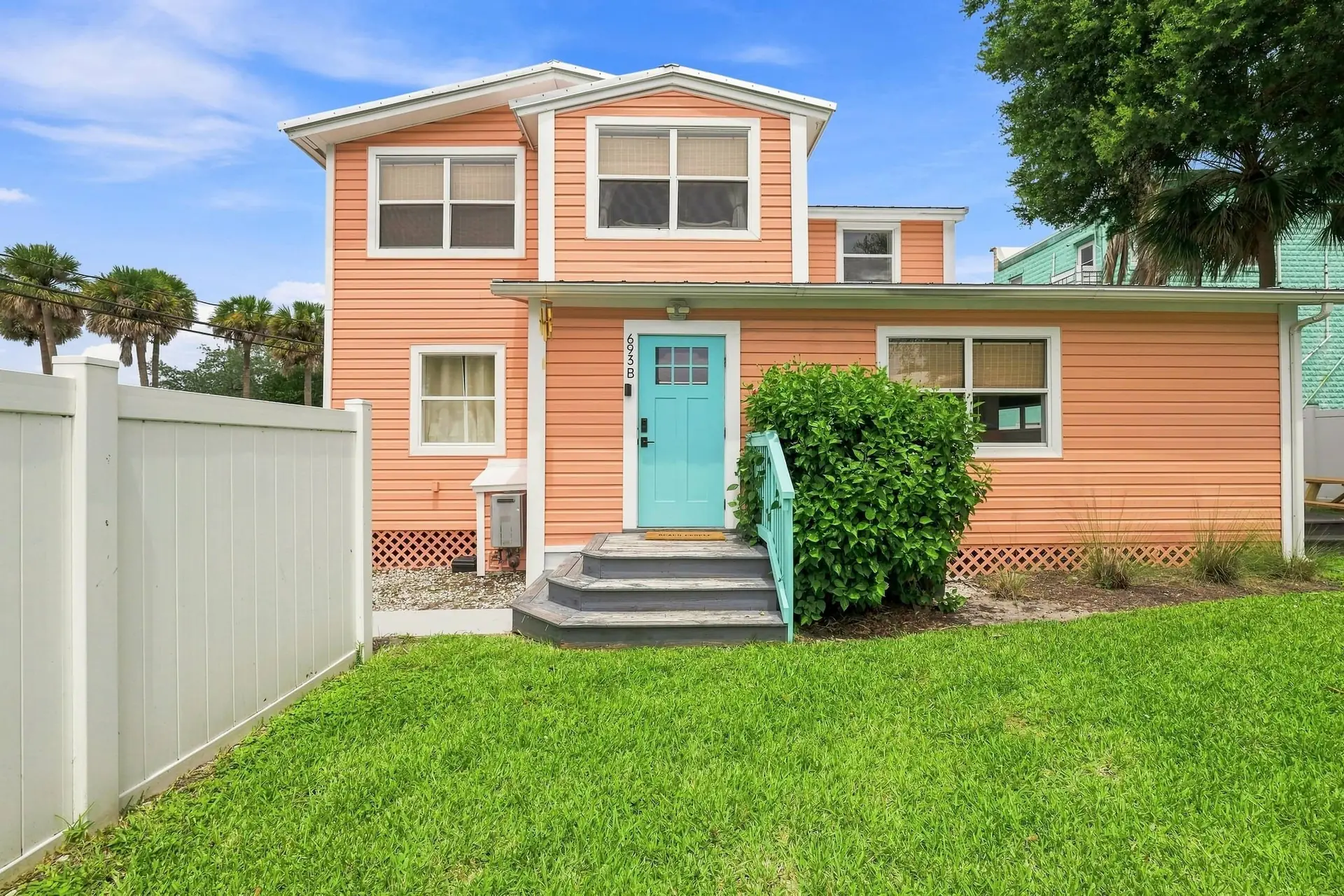 Front of the coral-painted coastal home with crisp white trim, a turquoise front door, wooden steps and a bright green lawn