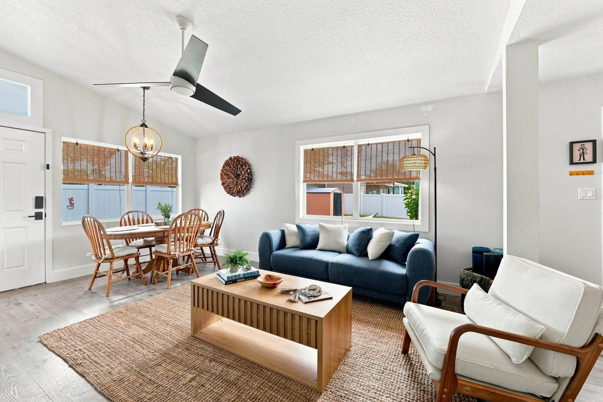 Vaulted living room looking toward the dining table and front door, with a ceiling fan, rattan globe chandelier and woven window shades