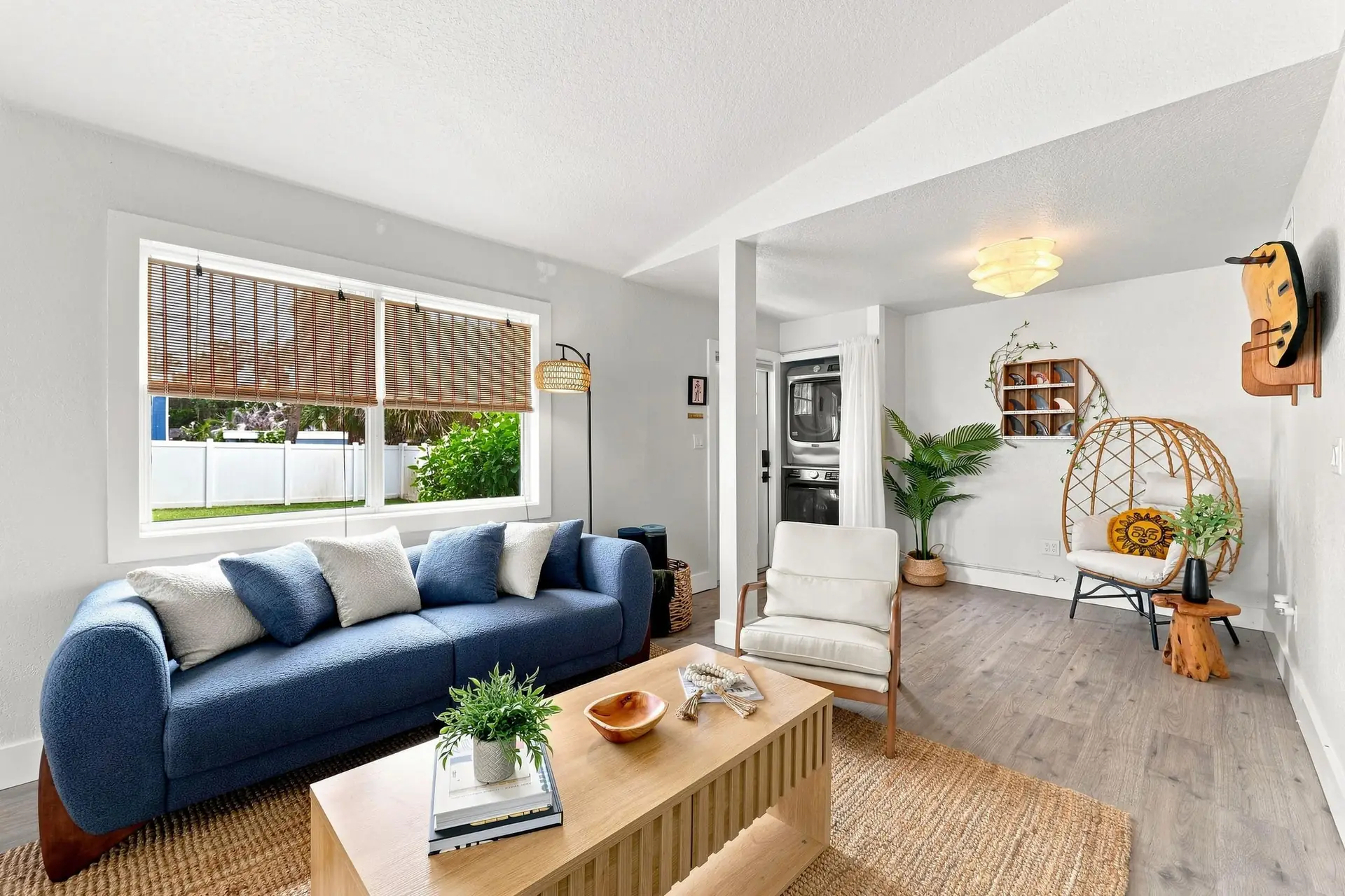 Living room with a blue bouclé sofa beneath a wide picture window with woven shades, a light oak coffee table on a jute rug, and a hanging rattan egg chair in the far corner