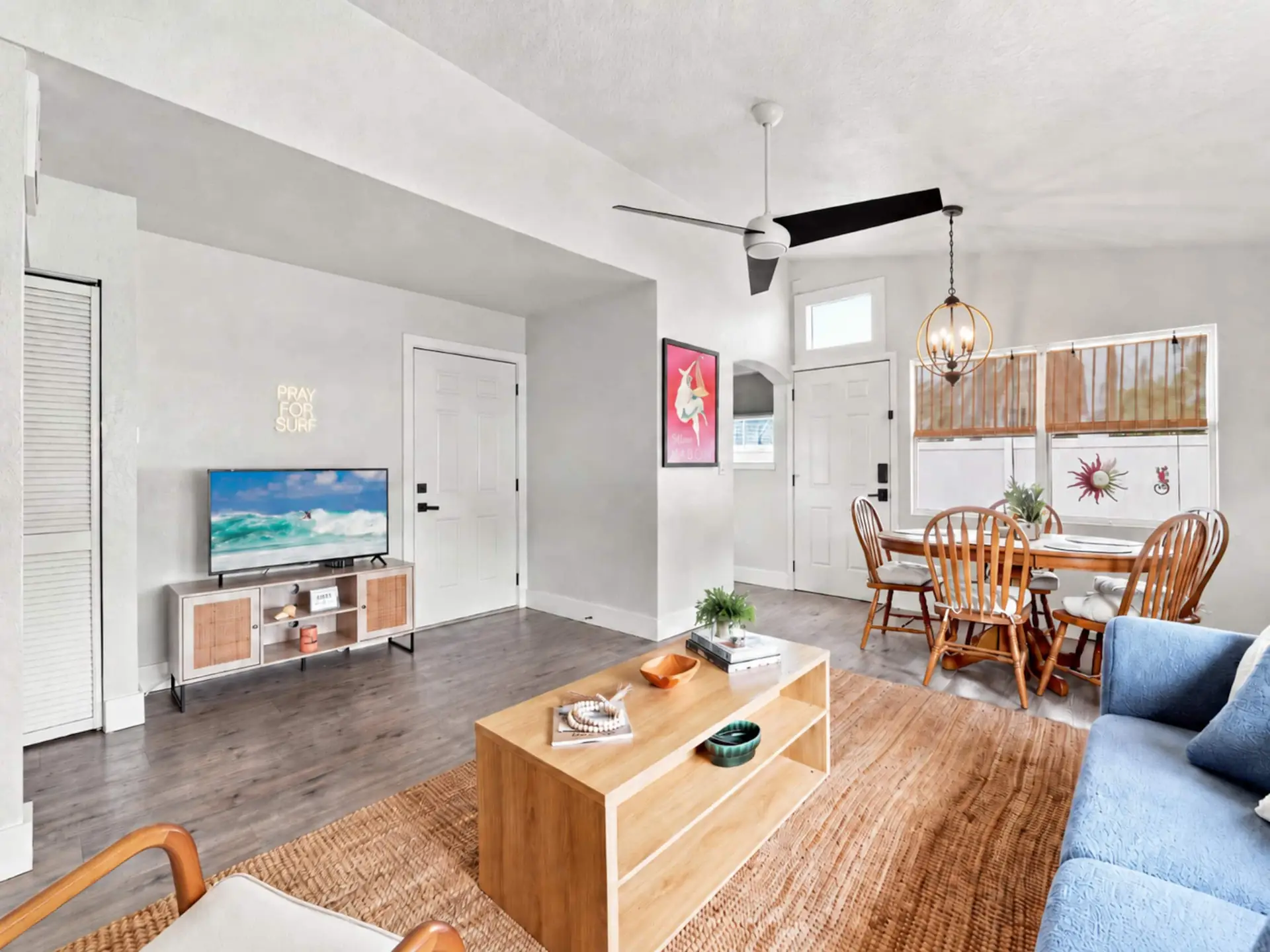 Open-plan living and dining area with a wall-mounted TV on a cane console, a "Pray for Surf" neon sign, and the dining table by the front door