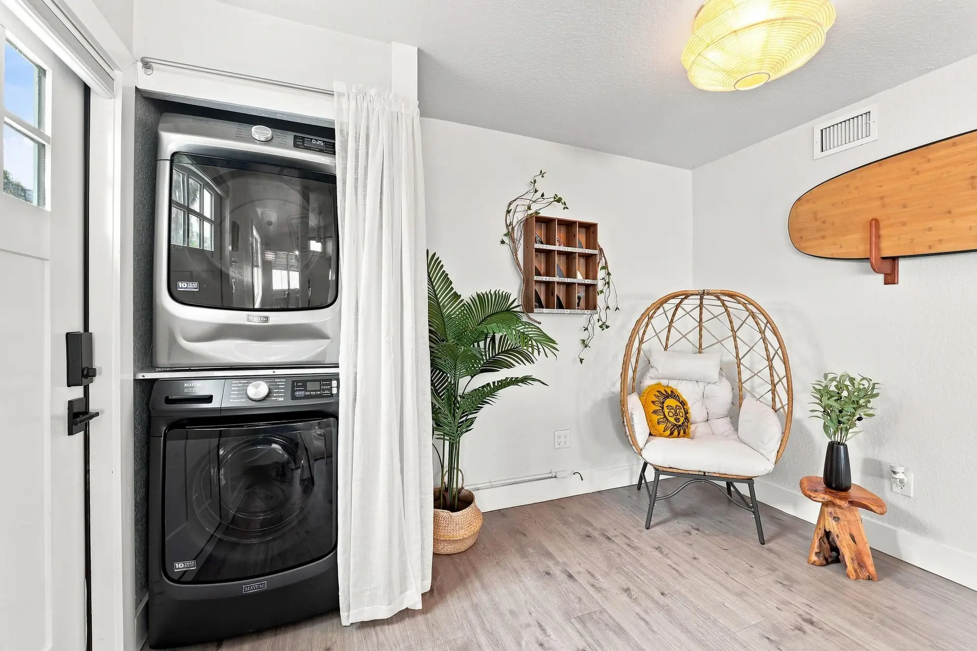 Cozy reading nook with a rattan egg chair and sun-embroidered pillow beside a full-size stacked washer and dryer, under a wooden surfboard on the wall